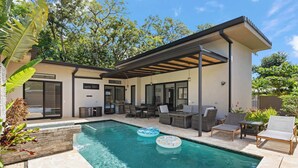 Un área de piscina moderna al aire libre con un patio cubierto, sillones de descanso y una zona de comedor.
