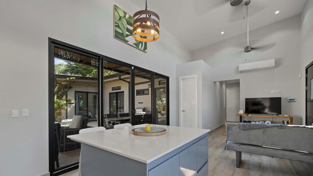 Una cocina moderna con una isla central, un comedor con puerta de cristal que da a un patio, y una sala de estar con sofá y televisor.