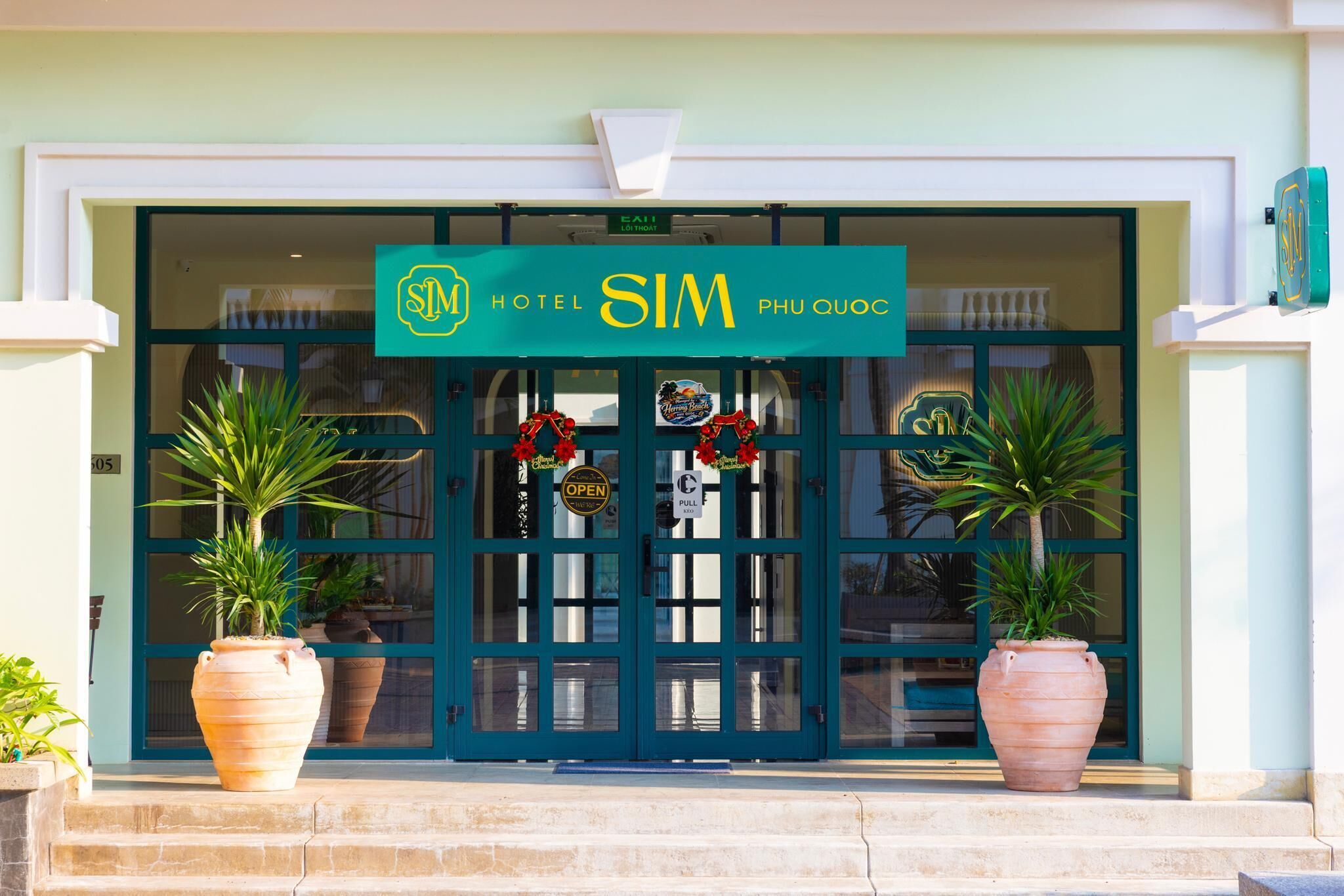 A hotel entrance with a green sign, double doors, and two large potted plants.