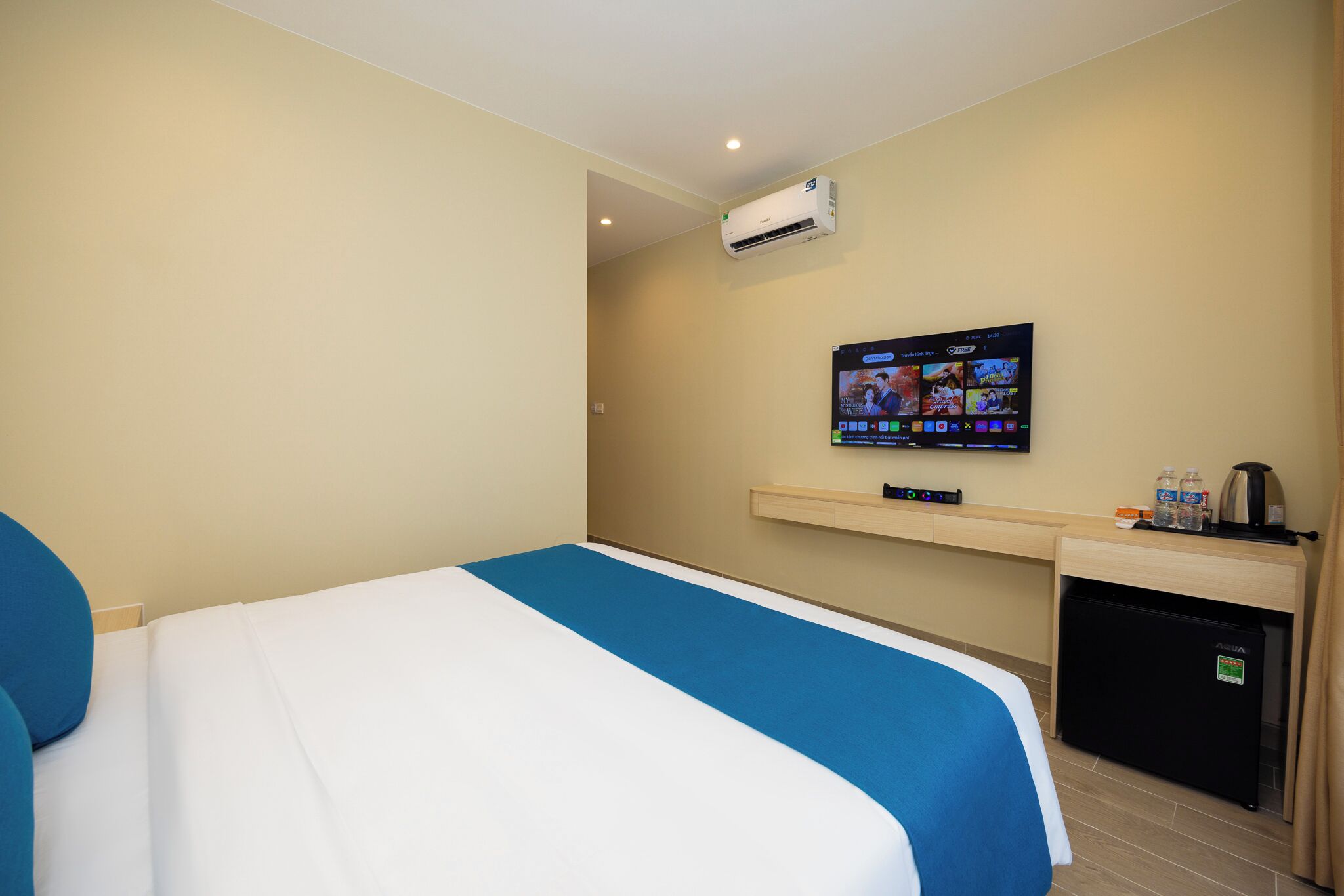 A hotel room with a bed, a TV mounted on the wall, a small desk with a coffee maker, and an air conditioning unit.