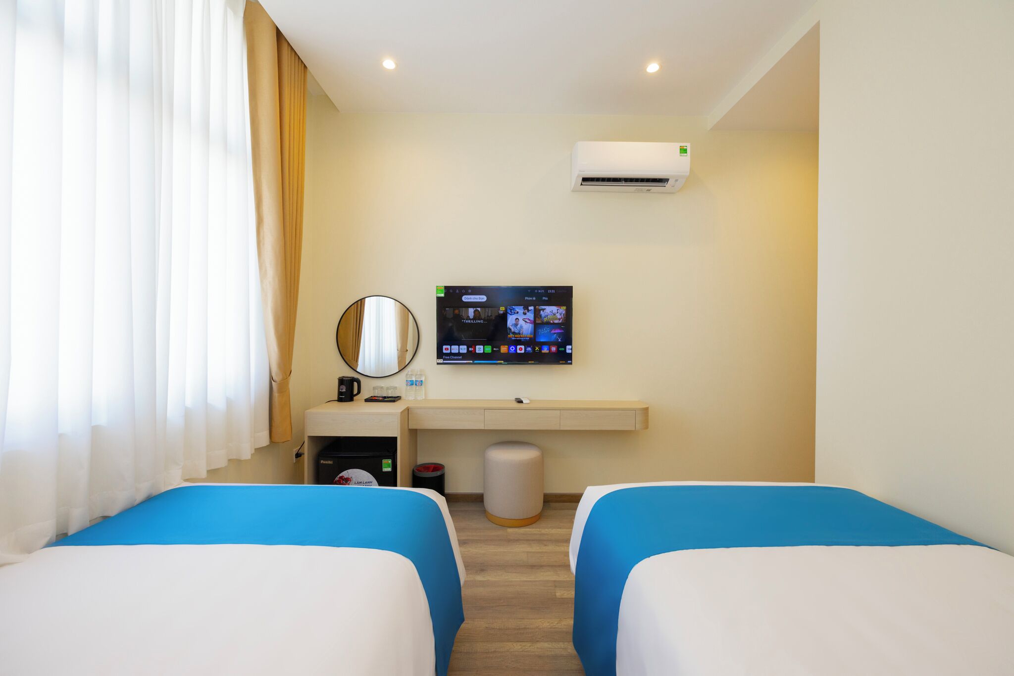 A hotel room with two beds, a TV, an air conditioner, a desk with a mirror, and a waste bin.