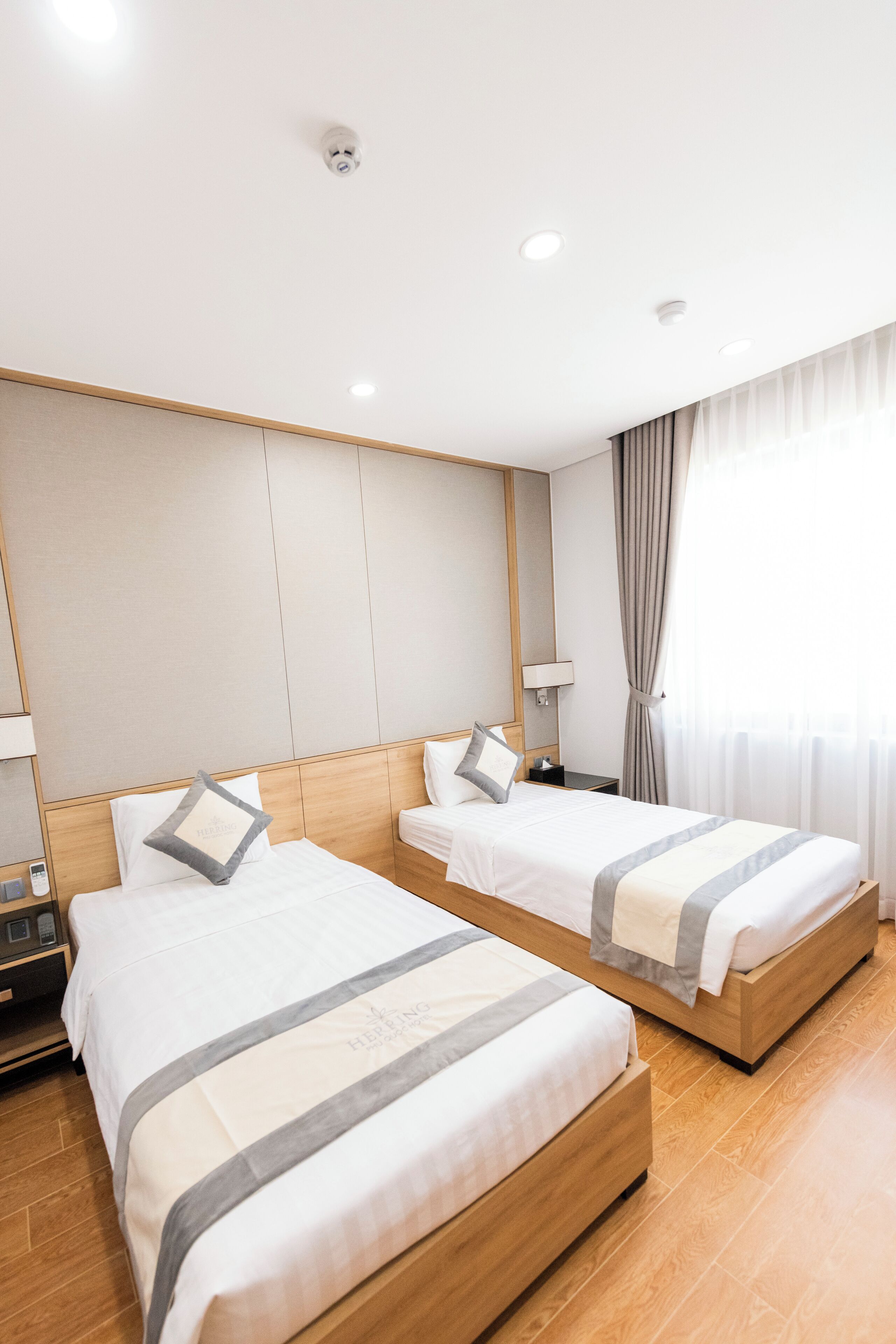 Habitación de hotel con dos camas, cabeceros de madera, una mesita de noche, una lámpara y una ventana con cortinas.