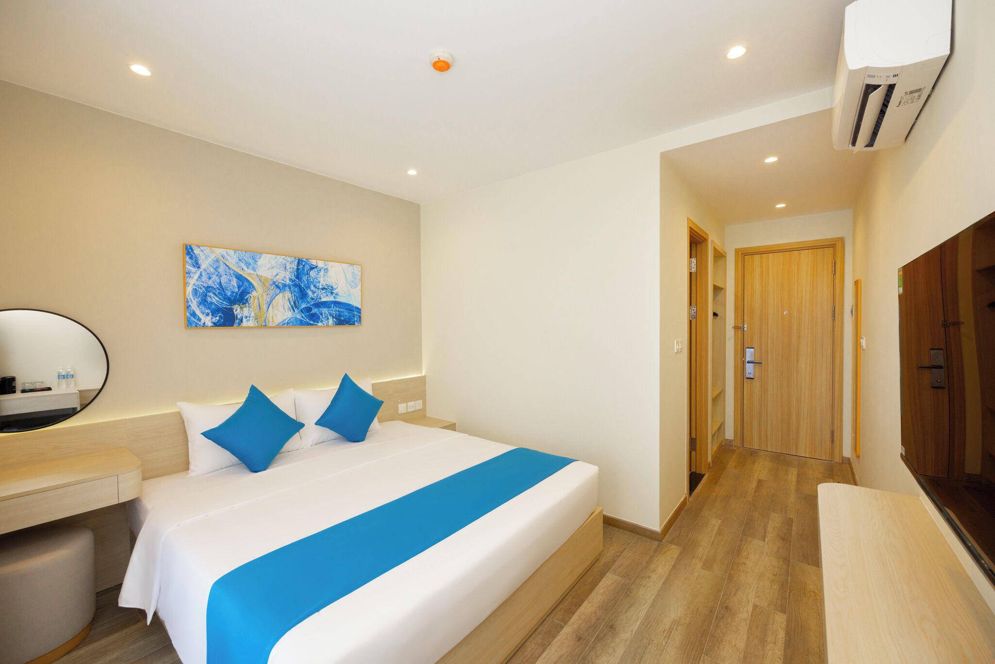 A hotel room with a bed, a desk, a mirror, a painting, and a wooden door.