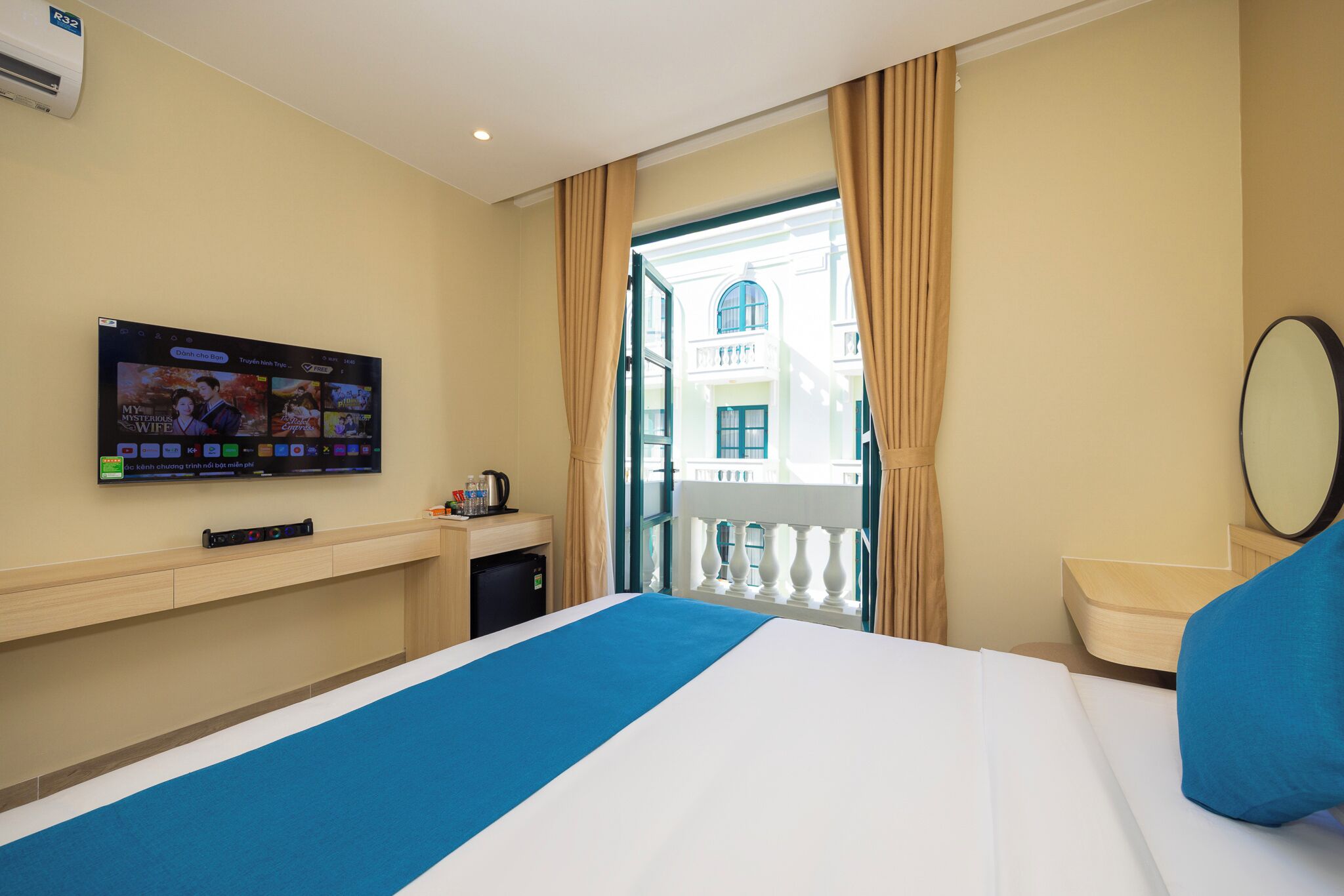 A hotel room with a bed, a TV mounted on the wall, a small table with a kettle, and a view of a balcony and building through the window.