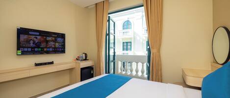 A hotel room with a bed, a TV mounted on the wall, a small table with a kettle, and a view of a balcony and building through the window.