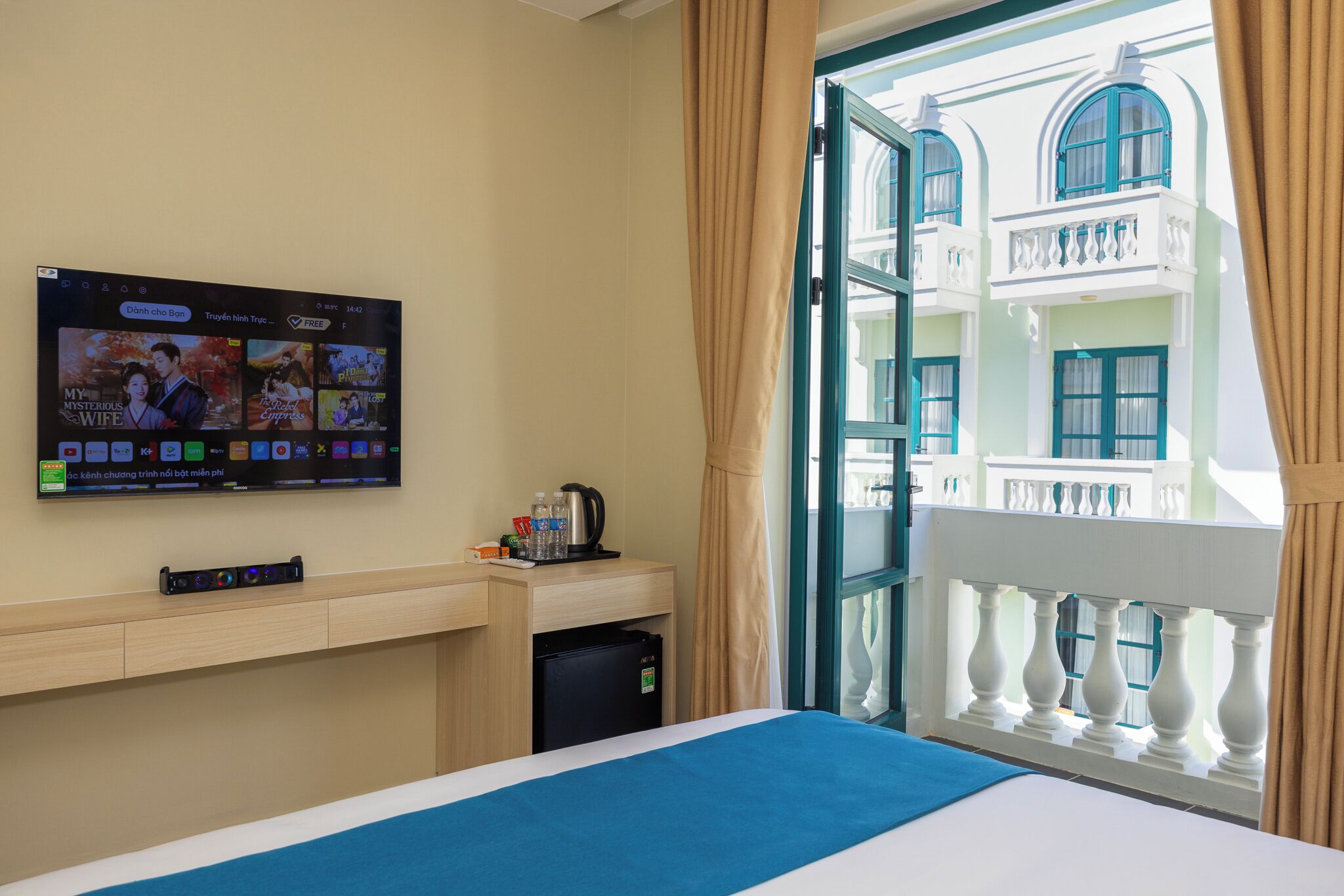A hotel room with a bed, a TV mounted on the wall displaying a movie, a small table with a kettle and cups, and a balcony with a view of a building.
