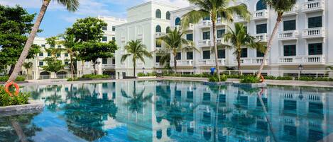 A swimming pool with clear blue water, surrounded by palm trees and a white building with balconies.