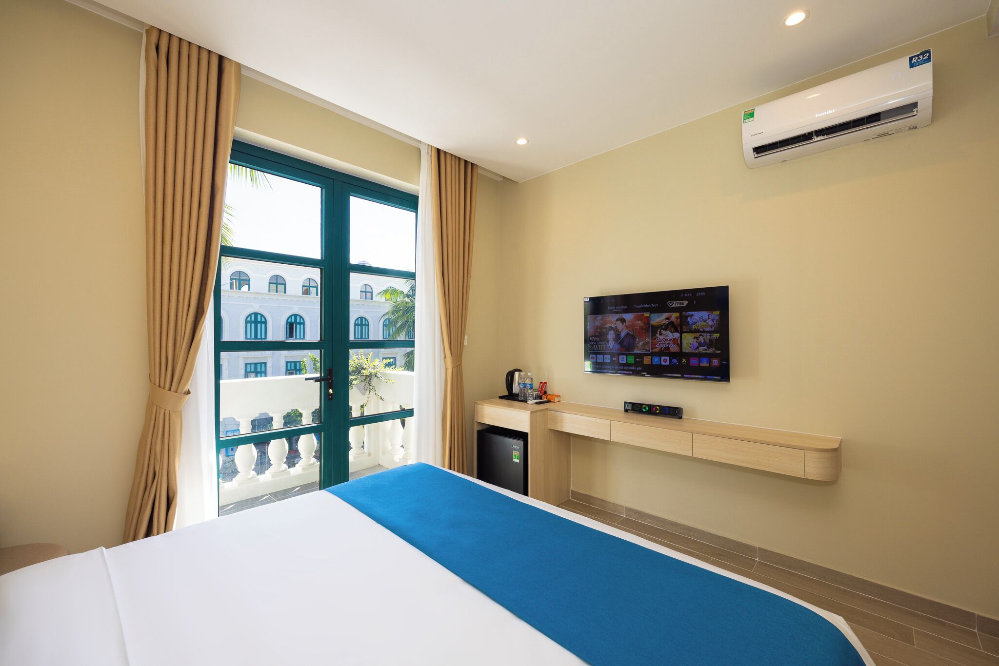 A hotel room with a bed, a TV on the wall, an air conditioner, and a view of buildings outside.