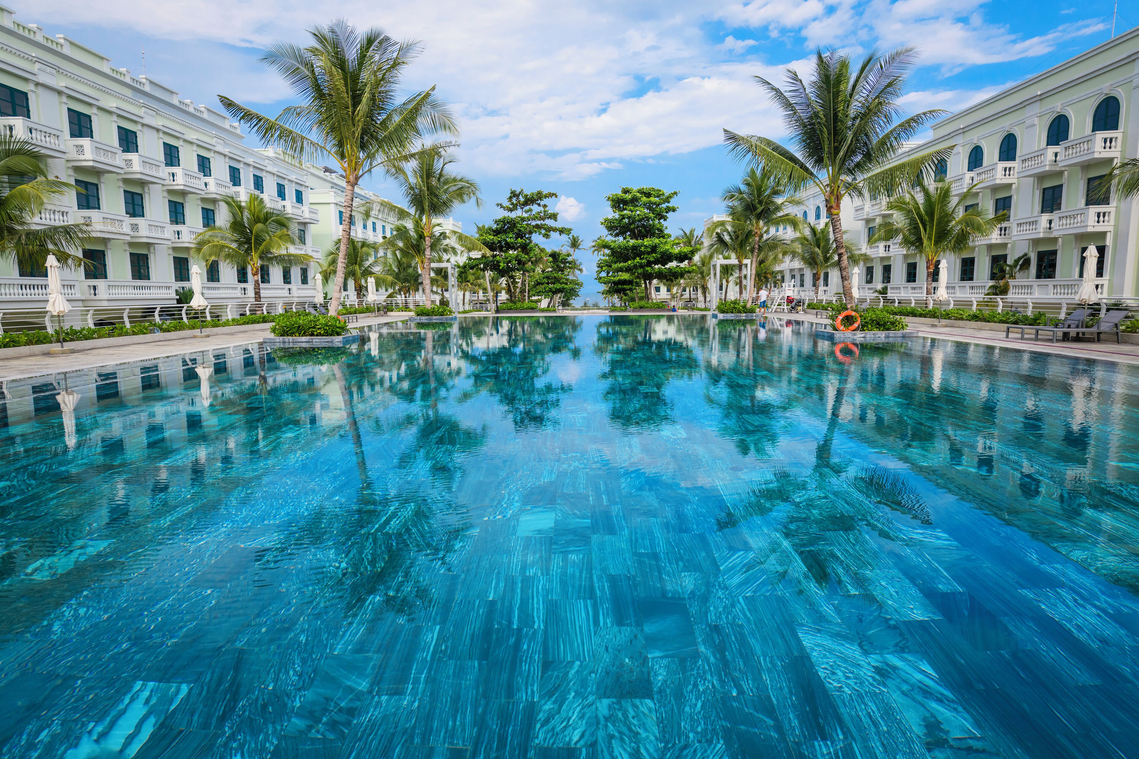 Une grande piscine aux eaux cristallines, entourée de bâtiments blancs dotés de balcons, de palmiers et surplombant un ciel dégagé.