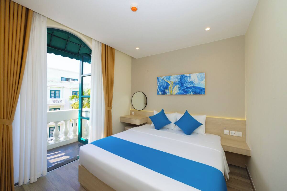 Une chambre d’hôtel comprenant un lit, une table de chevet, une commode, une fenêtre avec des rideaux, un tableau accroché au mur et une vue sur l’extérieur.