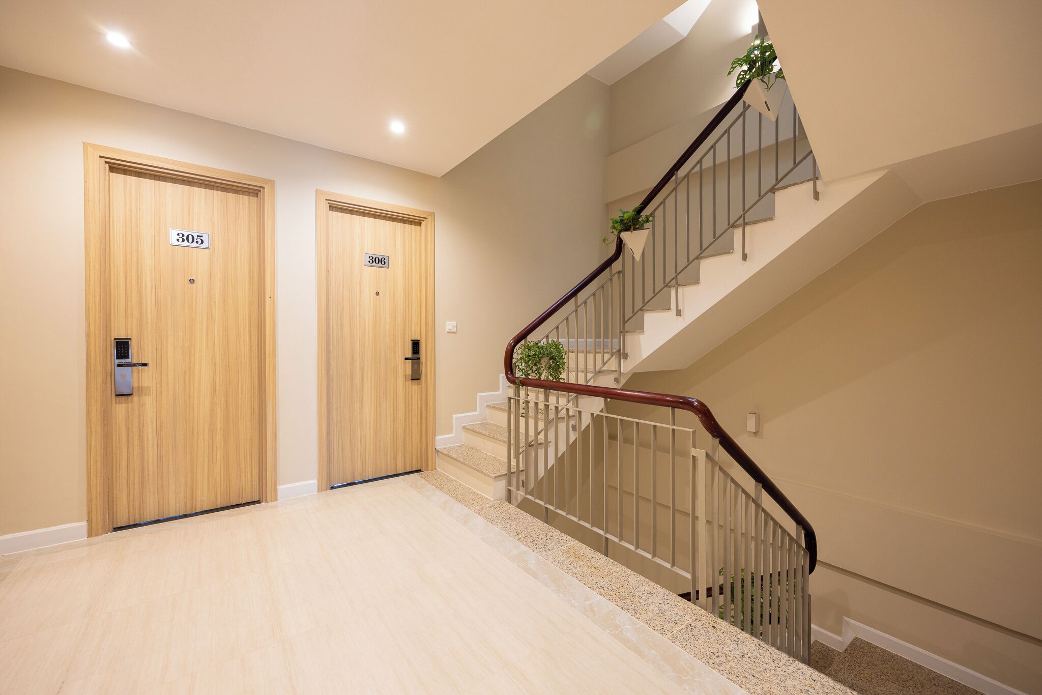 A modern hotel lobby with two wooden doors labeled 305 and 306, a staircase with a metal railing, and a plant on the railing.