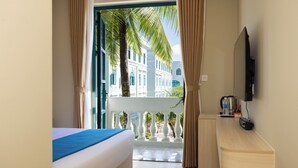 Hotelkamer met een bed, een klein bureau, een televisie en een balkon met uitzicht op gebouwen en palmbomen.