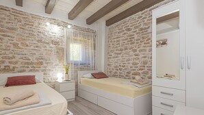 Une chambre avec un mur en pierre, un lit avec un matelas beige, une table de chevet, une armoire et une petite table.