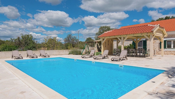 Une piscine avec des chaises longues, un espace de détente couvert et un bâtiment en pierre surmonté d’un toit de tuiles rouges.