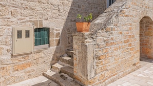 Un bâtiment en pierre avec un petit escalier menant à une porte, un pot de fleurs placé sur les marches et une fenêtre ornée d’une grille métallique.
