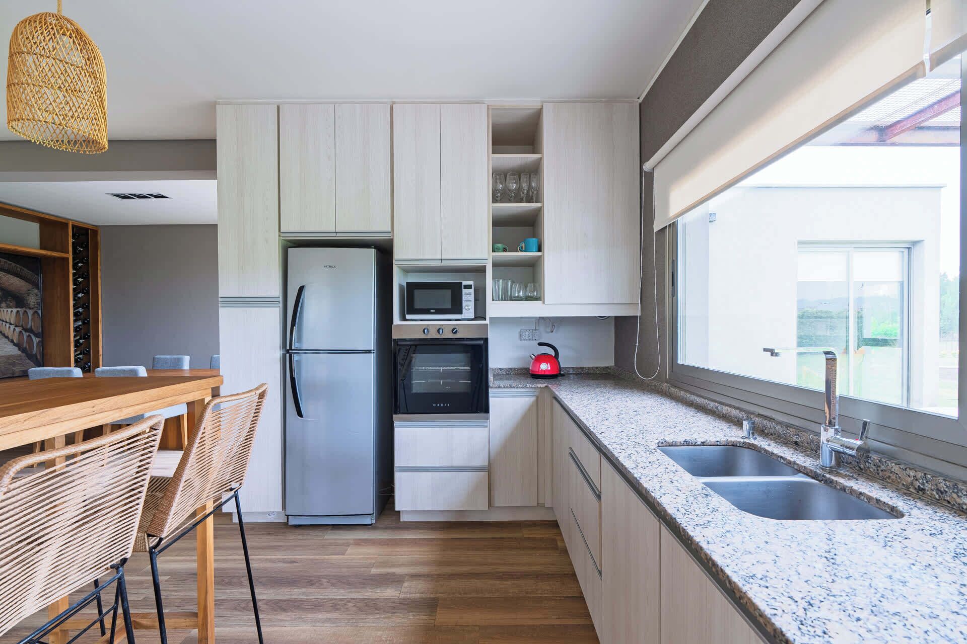 Cocina moderna con refrigerador, horno y microondas de acero inoxidable. Cuenta con un comedor con mesa de madera y sillas de mimbre. La cocina tiene un ventanal con vistas al exterior.