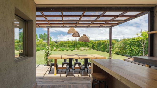 Un espacio exterior para comer con una mesa y sillas de madera, una pérgola con techo de malla y lámparas de mimbre colgantes.