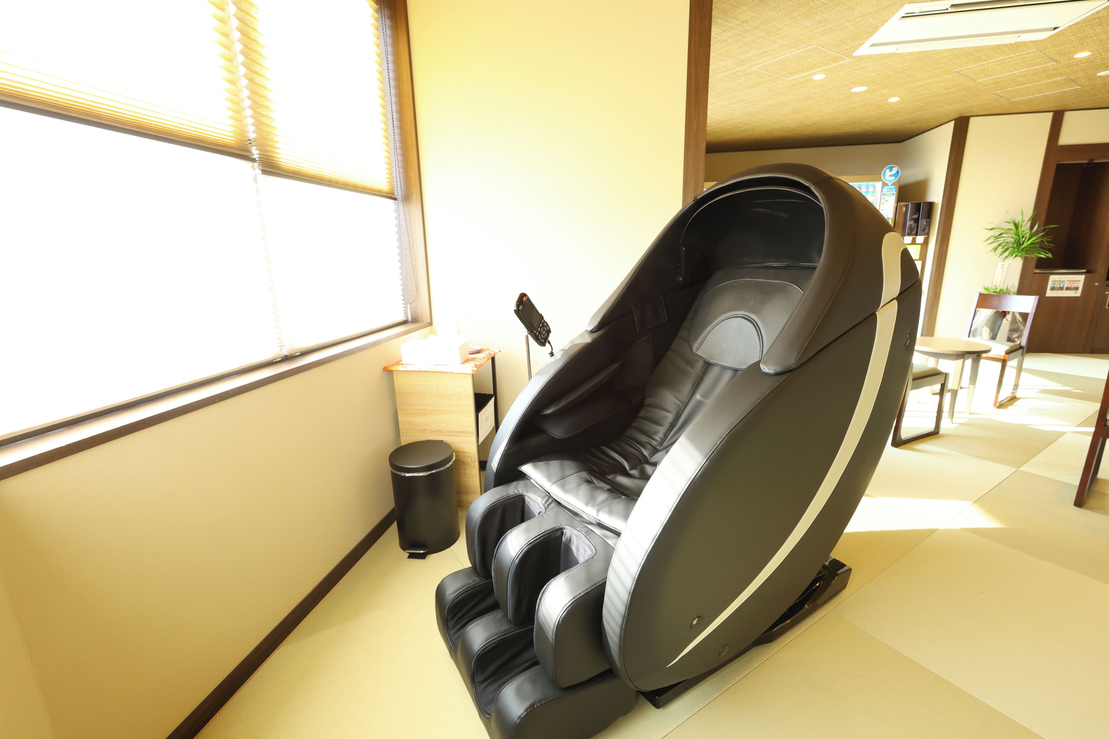 A massage chair in a room with a window, a trash bin, and a small table with a plant.