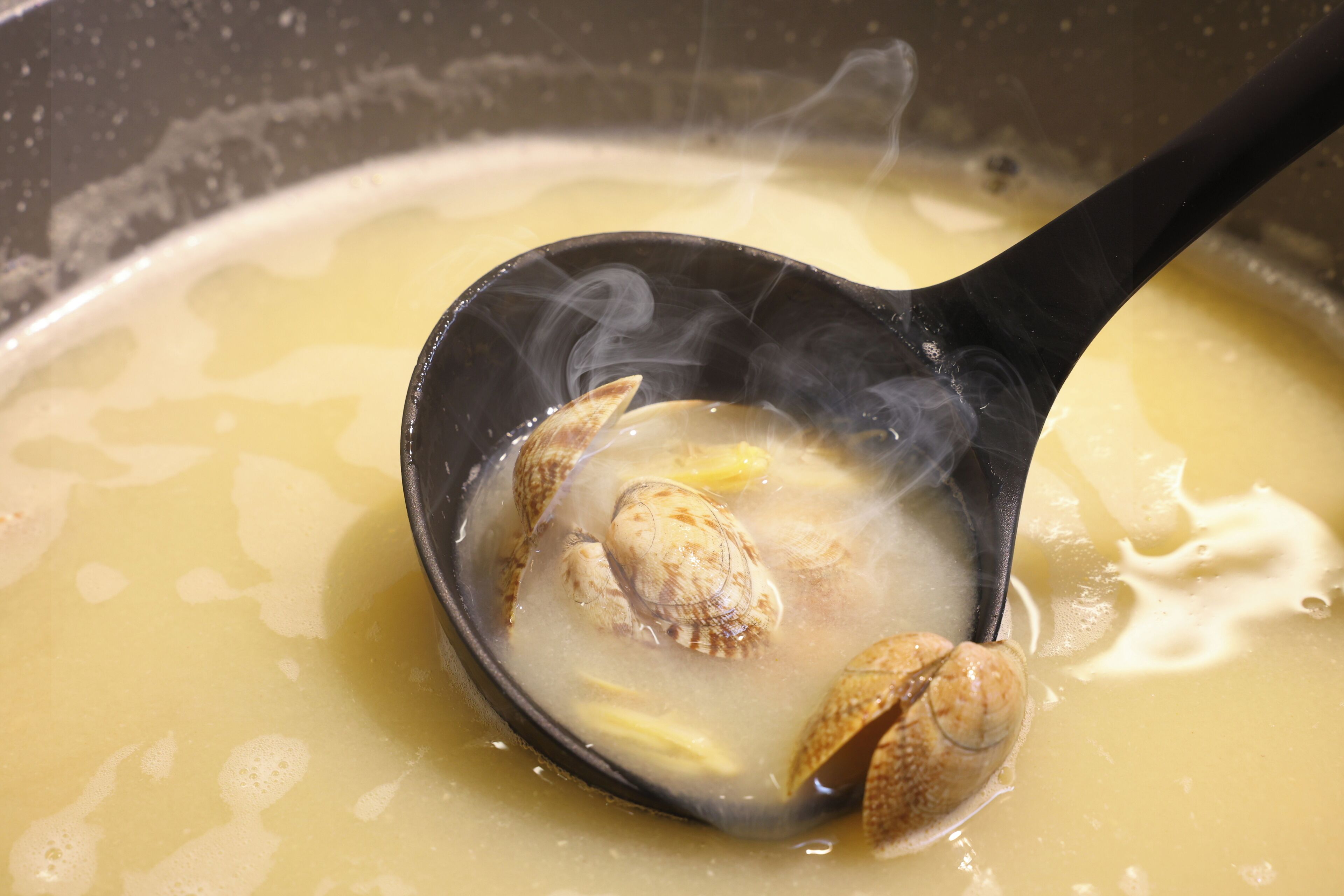 A close-up of a soup with clams in a black ladle.