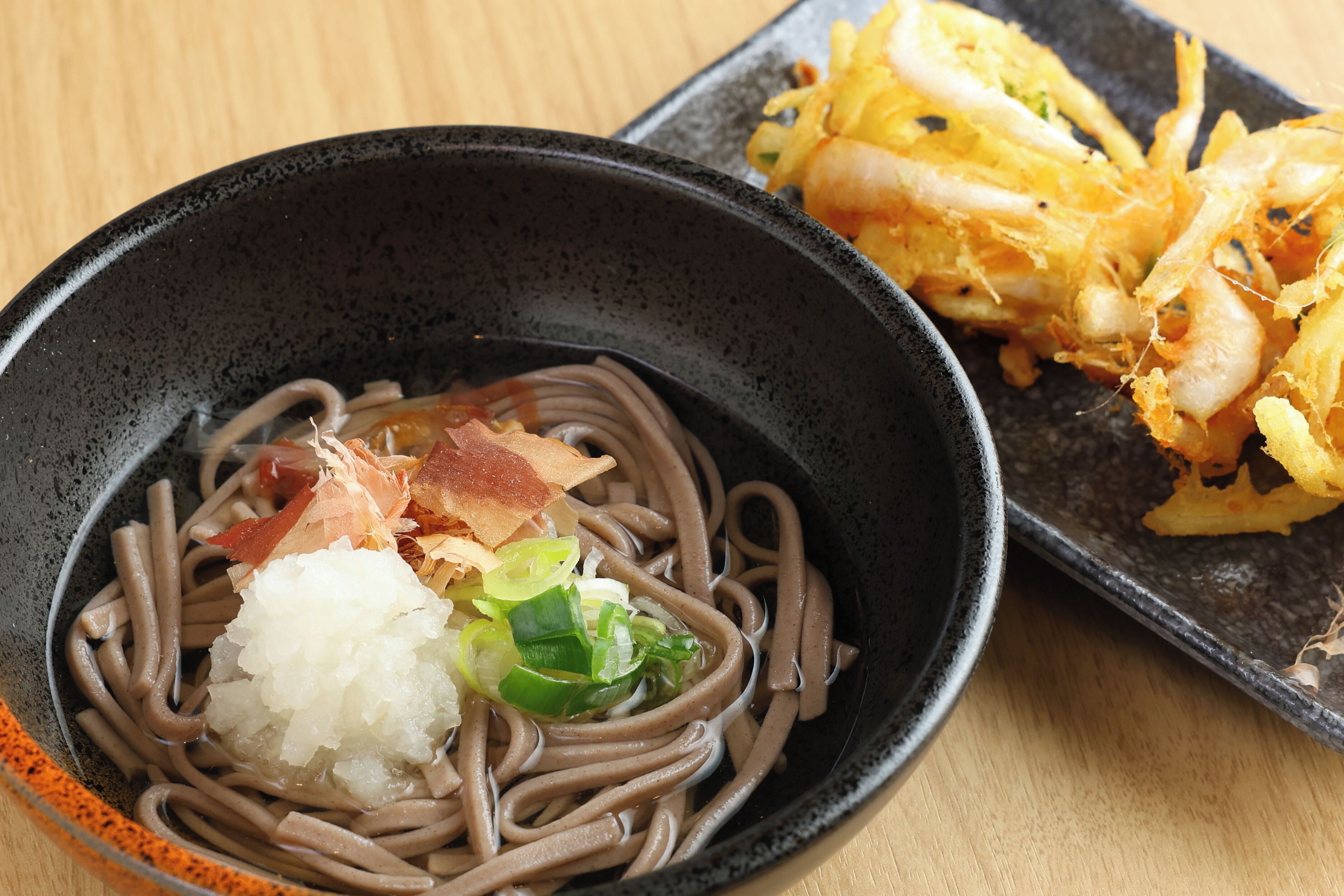 Un tazón de fideos soba con ingredientes, un plato de camarones tempura y una pequeña fuente de verduras encurtidas.