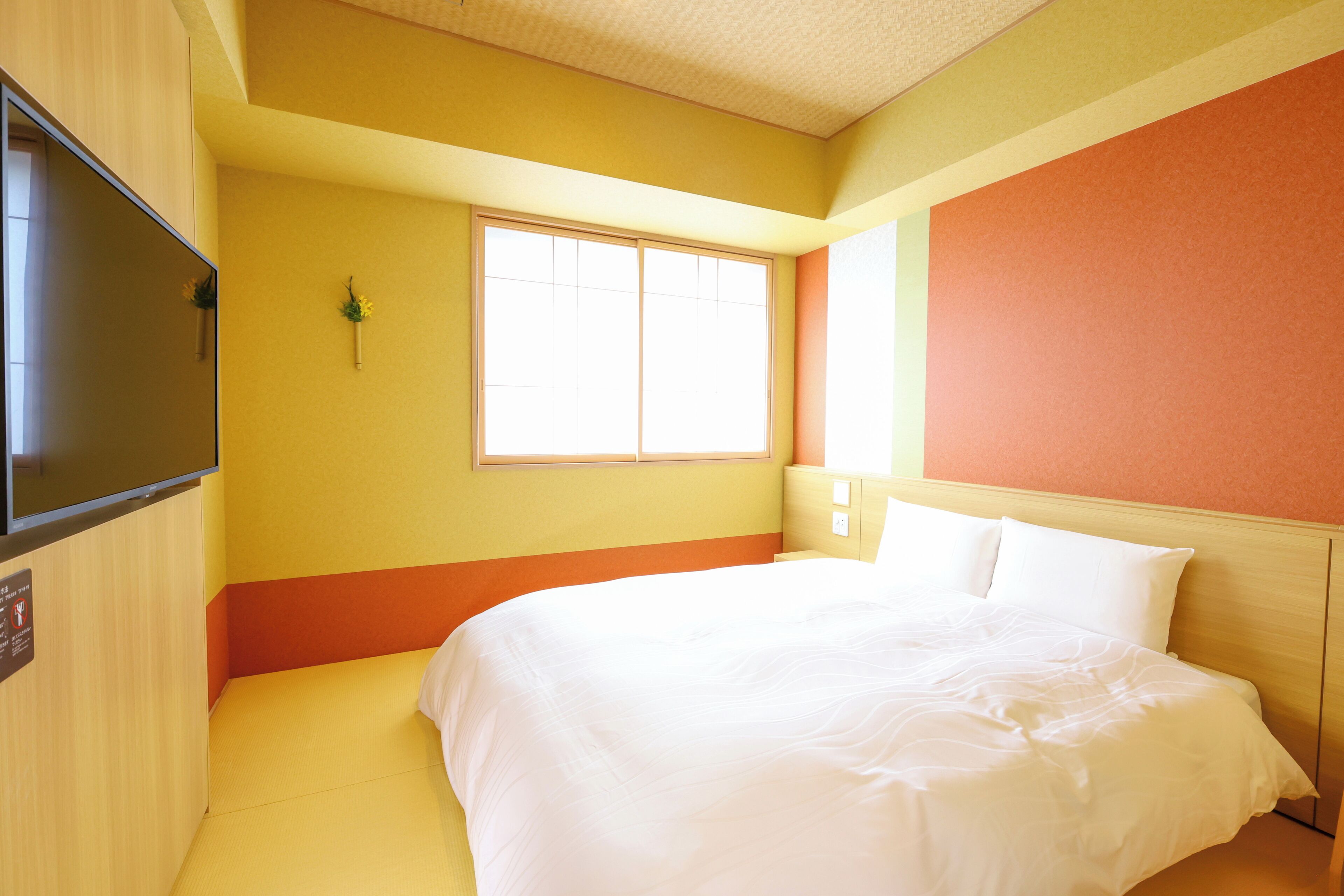 A hotel room with a bed, a television, a window, and a wall-mounted light fixture.