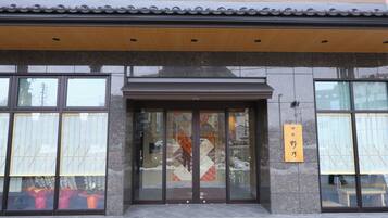 A building entrance with large glass doors, a wooden sign with Japanese characters, and a tiled roof.