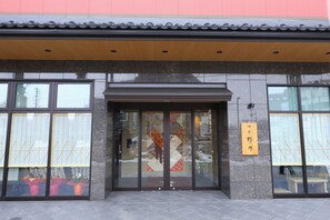 A building entrance with large glass doors, a wooden sign with Japanese characters, and a tiled roof.
