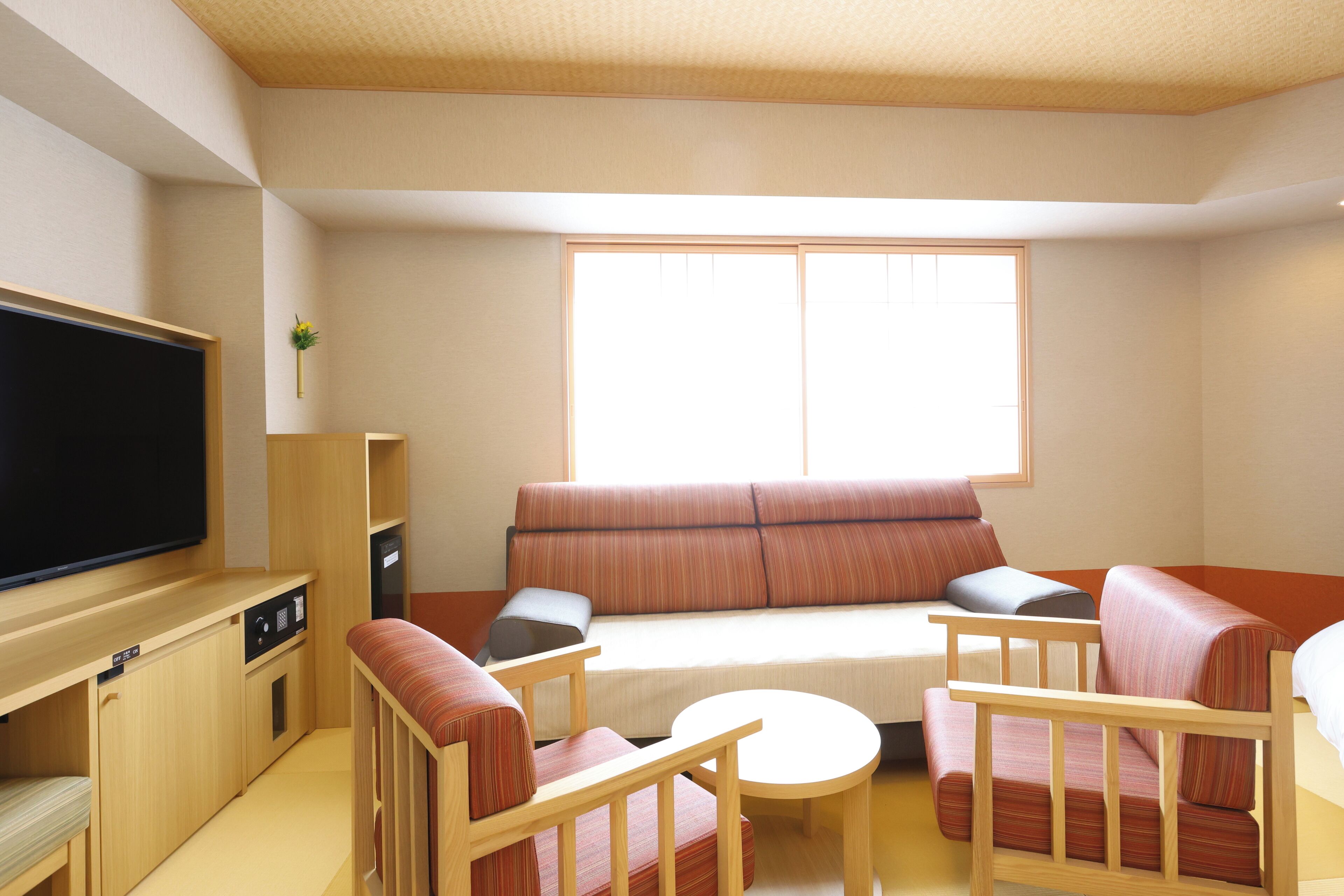 A compact living room with a sofa, two armchairs, a small table, a television, and a window providing natural light.