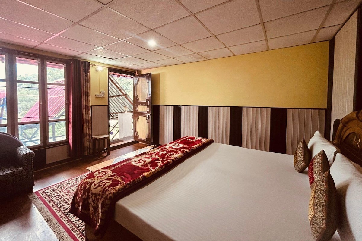Habitación de hotel con una cama grande, cabecera de madera, colcha de rayas rojas y blancas, ventana con vistas a un edificio de techo rojo y una puerta que da al exterior.