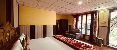 Habitación de hotel con una cama grande, cabecera de madera, una silla, un armario y una ventana con cortinas.
