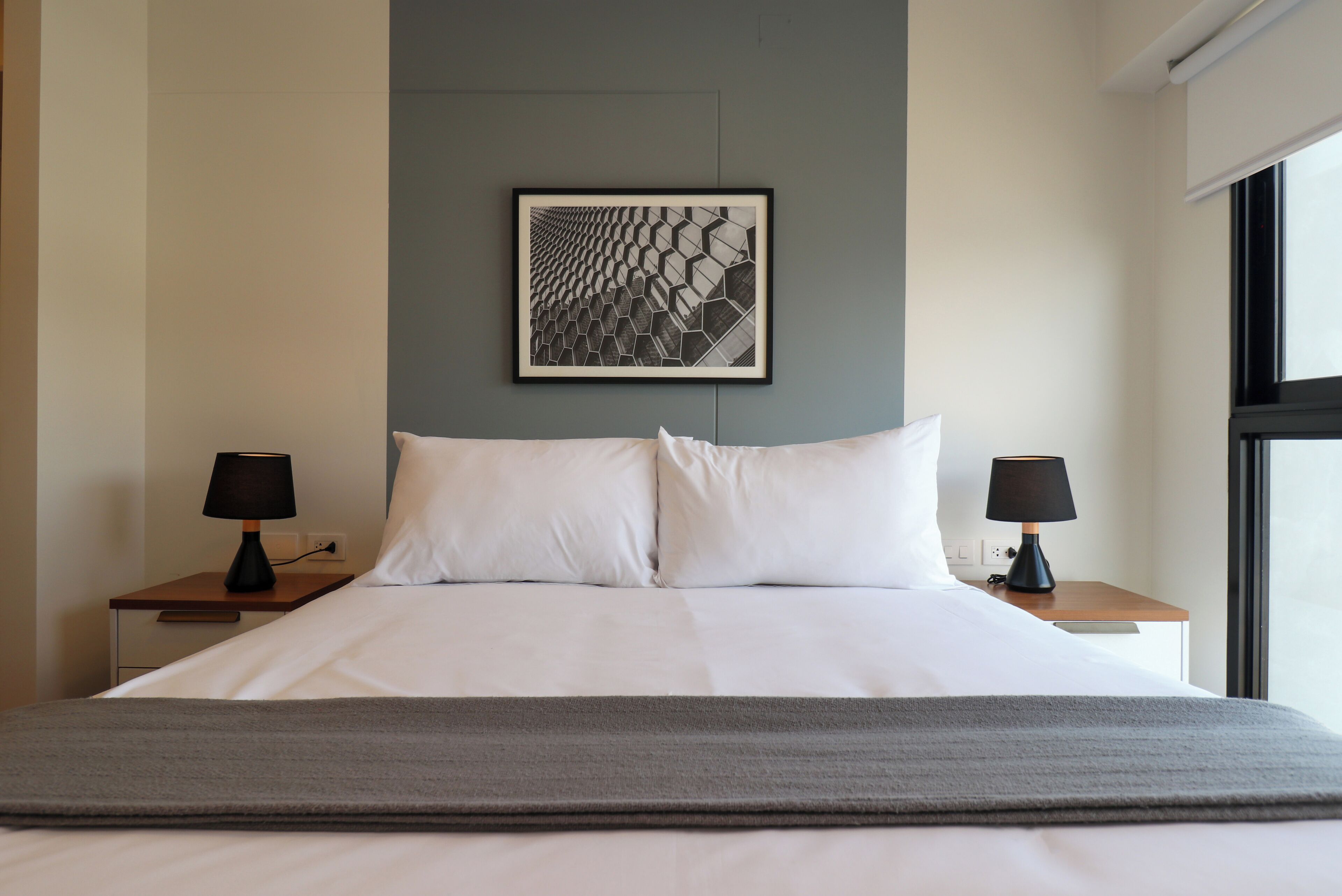 A neatly made bed with white linens, two bedside tables with lamps, a framed artwork on the wall, and a window on the right.