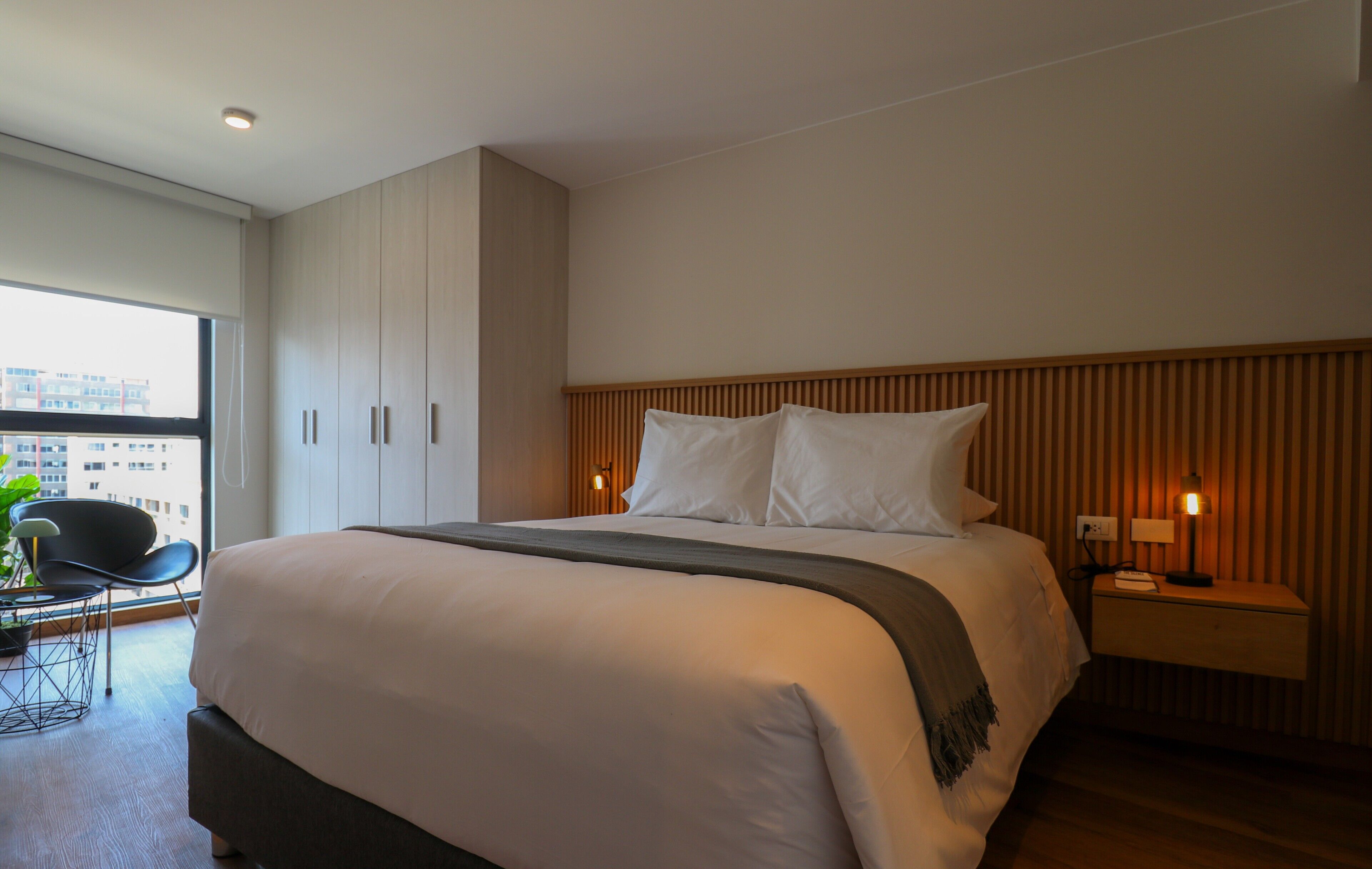A modern hotel room with a large bed, a nightstand with a lamp, a chair, and a view of the city through a large window.