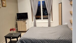 Une chambre moderne avec un lit, une télévision, une petite table avec un vase et une fenêtre avec des rideaux.