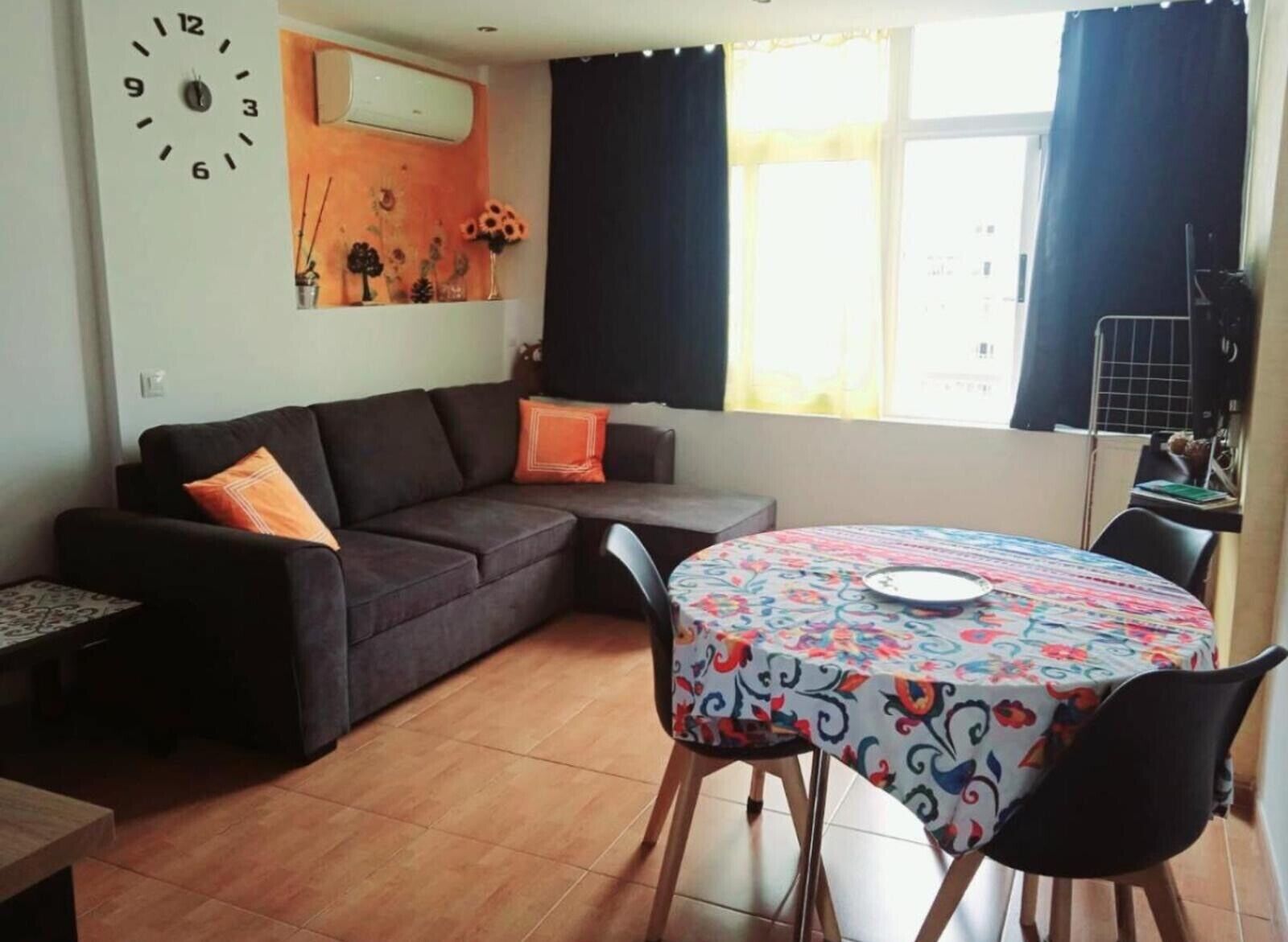 Ein Wohnzimmer mit einer Couch, einem runden Tisch mit Stühlen, einer Uhr und einem Fenster mit Vorhängen.