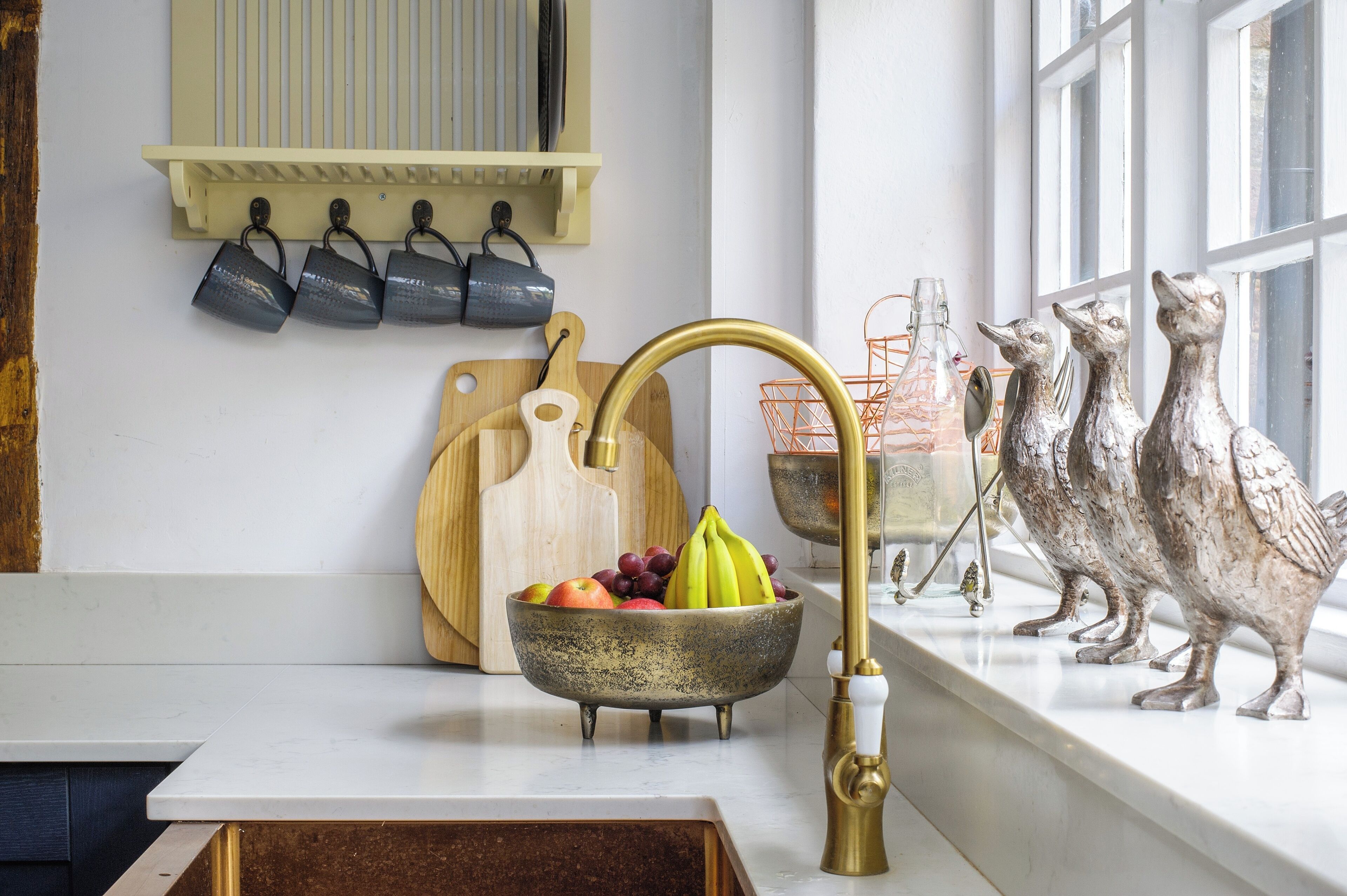 A kitchen with a white countertop, a brass faucet, a fruit bowl, a wooden cutting board, and decorative bird figurines.