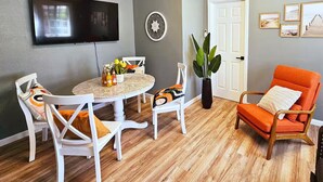 A modern living room with a round table, white chairs, a flat-screen TV, a decorative wall piece, and a door with framed pictures. - Bright Bungalow steps from Caltech & PCC, Close to the Rose Bowl, Kingsize Bed! (Pasadena)