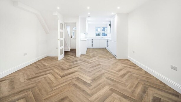 Amplia habitación con suelo de madera en espiga, paredes blancas y una cocina visible a través de una puerta abierta.