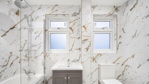 Un baño con paredes de mármol, una bañera, un lavamanos, un inodoro y dos ventanas.