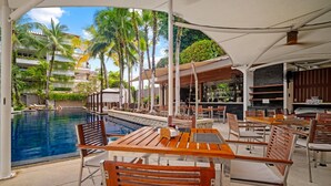 Een buitenzwembad met houten tafels en stoelen, omringd door palmbomen en een bar.