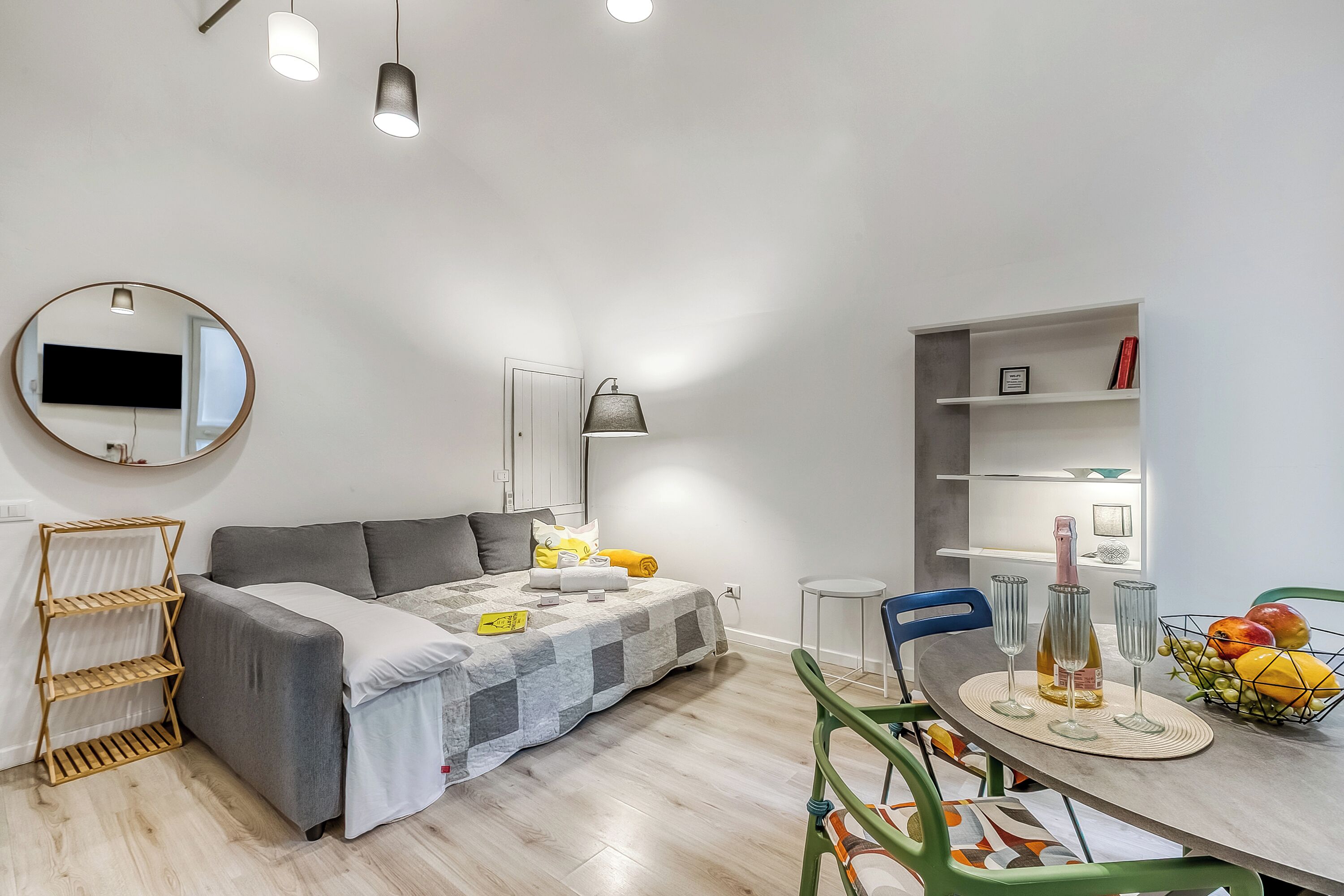 A modern living room with a sofa, a dining table with chairs, a fruit basket, and a TV mounted on the wall.
