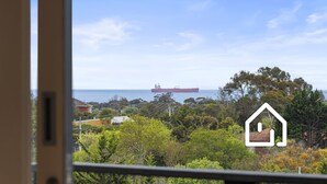 Blick aus einem Fenster auf ein großes, rotes Schiff auf dem Meer, mit Blick auf eine Wohngegend mit Bäumen und Häusern.