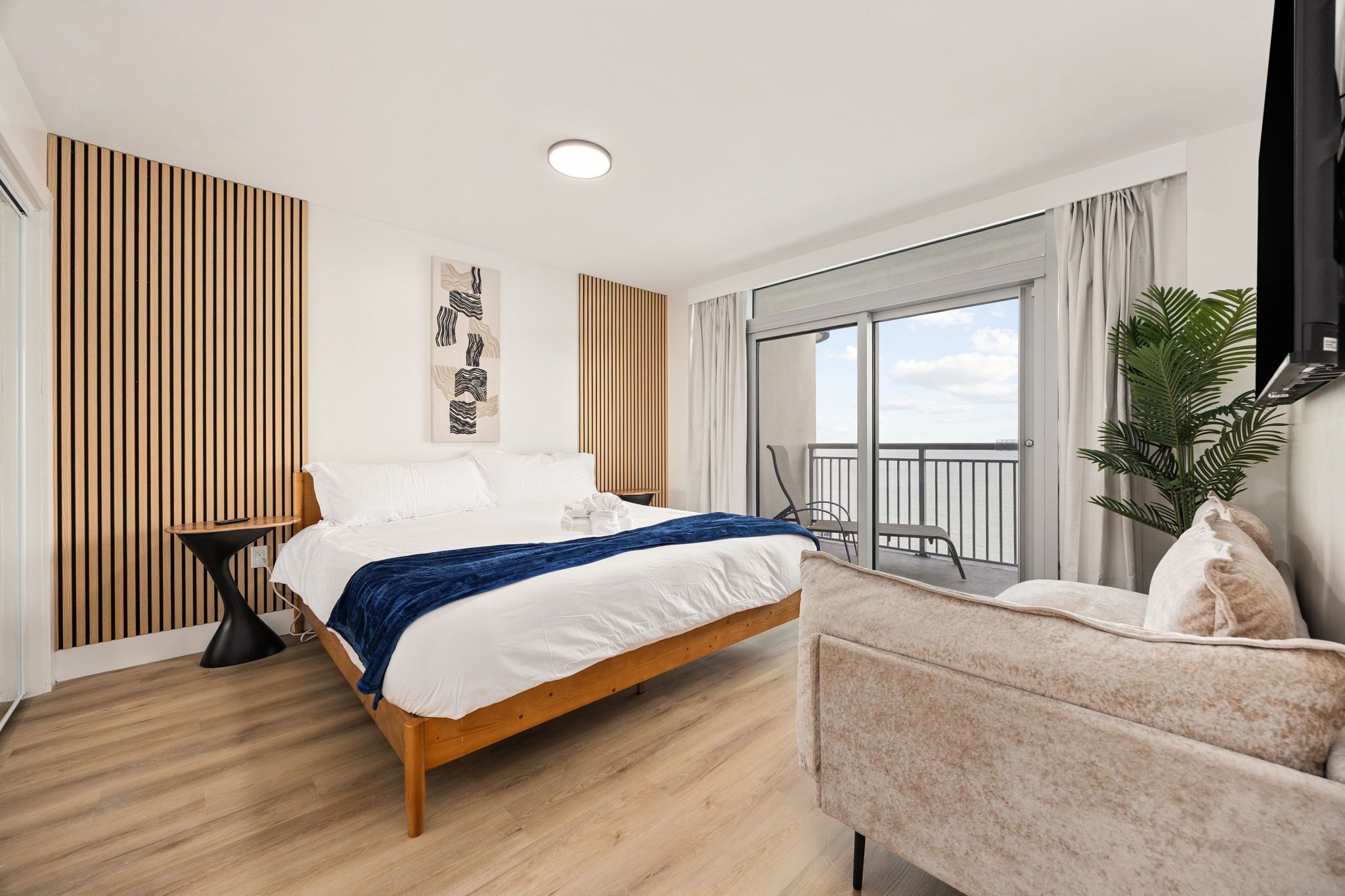 Una habitación de hotel moderna con una cama de madera, una mesita auxiliar pequeña, un sofá, un ventanal con balcón y un televisor de pantalla plana montado en la pared.