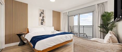 Una habitación de hotel moderna con una cama de madera, una mesita auxiliar pequeña, un sofá, un ventanal con balcón y un televisor de pantalla plana montado en la pared.