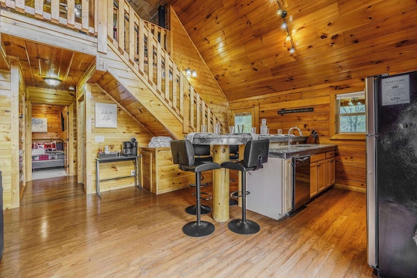 Interior de una cabaña de madera con cocina, comedor y una escalera que sube a un altillo.