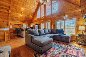 Interior acogedor de una cabaña con un techo de madera grande, un sofá modular, una zona de comedor y ventanas amplias.