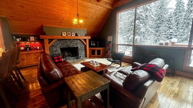 A cozy cabin interior with a stone fireplace, wooden ceiling, and a large window overlooking a snowy forest. - Creekside Family Hideout @ KHMR - Ski in/Ski Out (Golden)