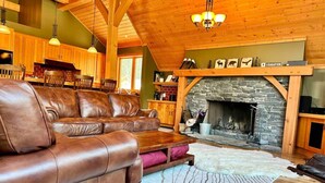 A cozy living room with a leather sofa, a stone fireplace, wooden beams, and a dining area. - Creekside Family Hideout @ KHMR - Ski in/Ski Out (Golden)
