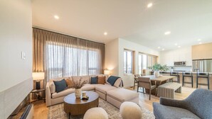 Een moderne woonkamer met een bank, salontafel en voetenbankje, een eethoek met stoelen, en een keuken met een koelkast en magnetron.