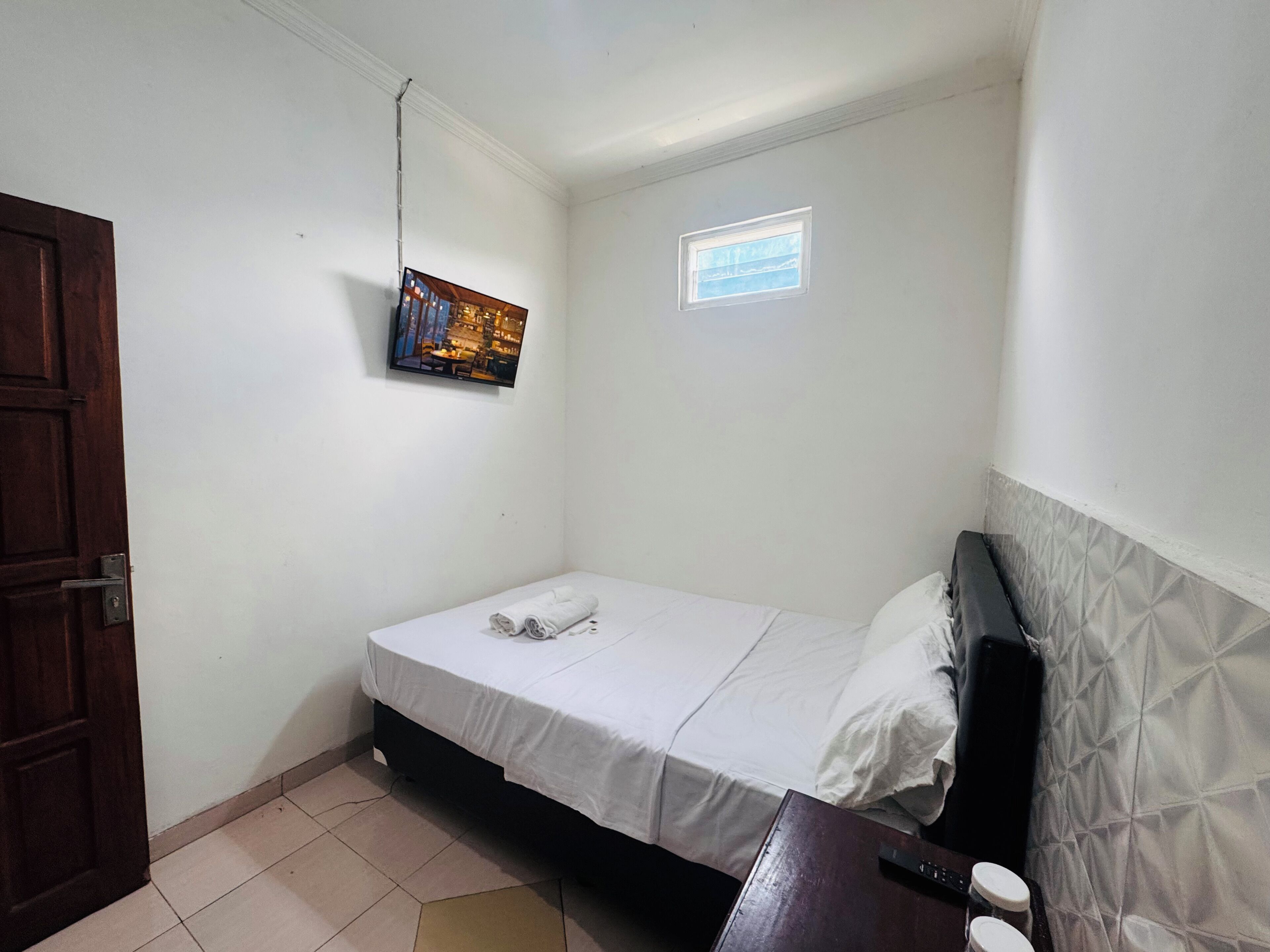 A small, clean room with a single bed, a wooden door, a small window, and a wall-mounted TV.