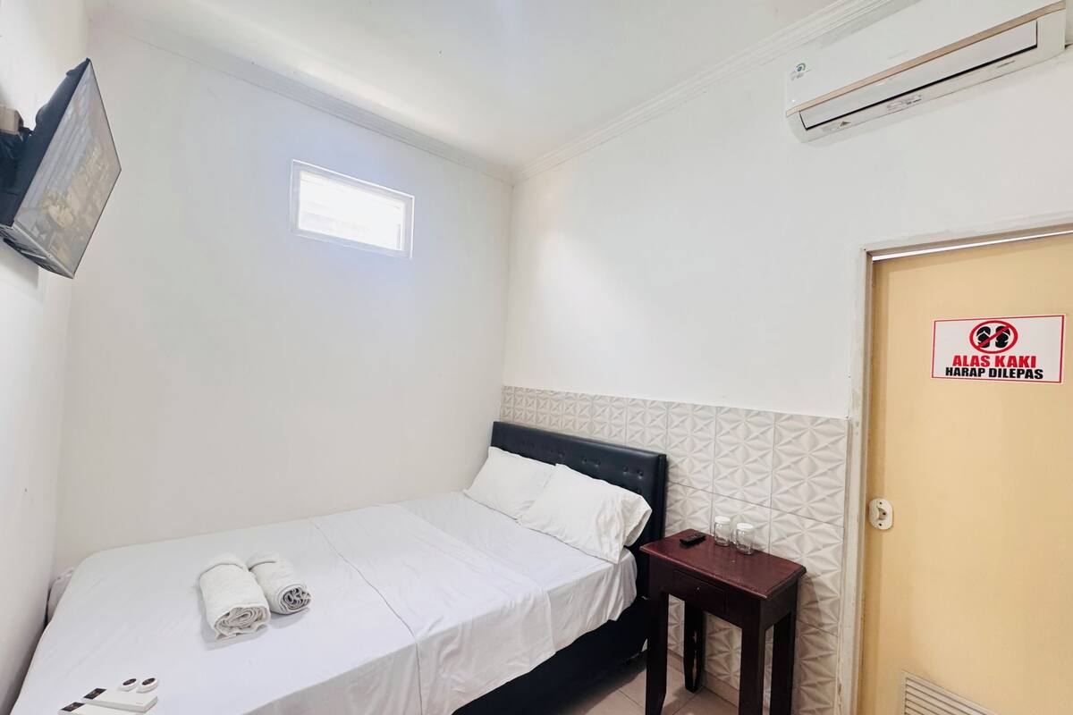 Una cama bien hecha con ropa blanca, una mesita de noche con un control remoto y vasos, un aire acondicionado de pared y una puerta con un letrero.